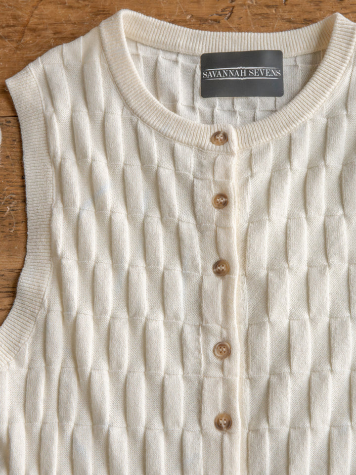 The Patero by Savannah Sevens western life{&}style is a cream-colored sleeveless top with a textured knit pattern and button-up front, shown laid flat on a wooden surface.