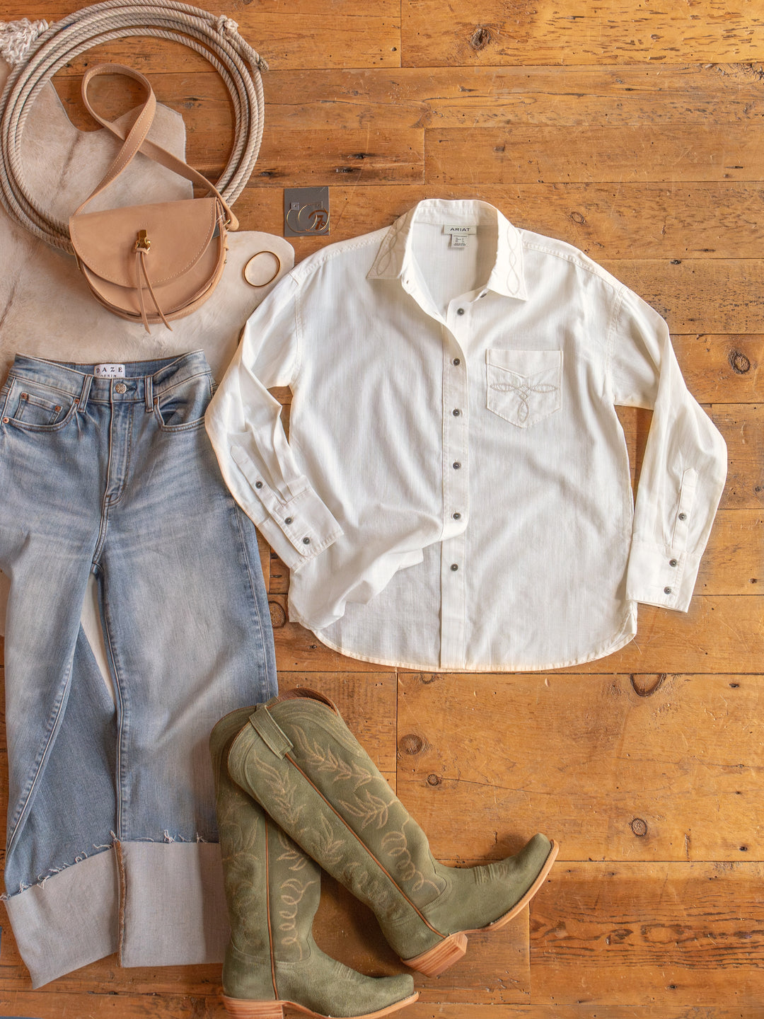 Flat lay: The Lawler off-white button up by Savannah Sevens western life{&}style (S-XXL), with an embroidered collar, light blue jeans, green cowboy boots, tan crossbody bag, beige scarf, and gold bangle on a wooden floor.