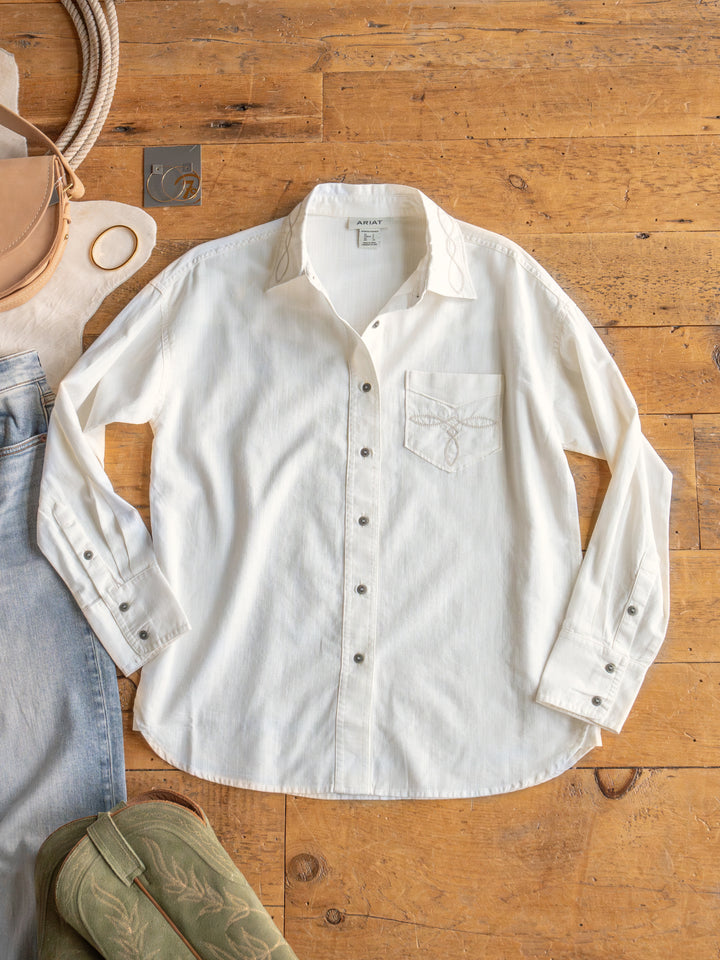 The Lawler by Savannah Sevens western life{&}style, an off-white button up with embroidered collar and front pocket, is laid flat on a wooden floor surrounded by a tan bag, light blue jeans, and green cowboy boots. 