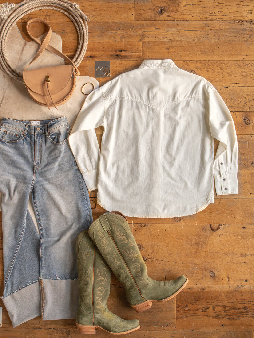 Flat lay of a casual outfit on a wooden floor, featuring The Lawler button up top by Savannah Sevens western life{&}style, light blue jeans, green cowboy boots, a tan crossbody bag, and accessories like a bracelet and ring.