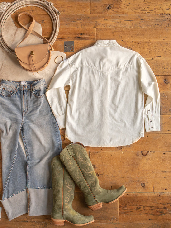 Flat lay of a casual outfit on a wooden floor, featuring The Lawler button up top by Savannah Sevens western life{&}style, light blue jeans, green cowboy boots, a tan crossbody bag, and accessories like a bracelet and ring.