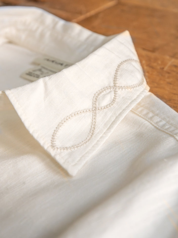 A close-up of The Lawler button up top by Savannah Sevens western life{&}style, featuring an off-white western yoke and embroidered infinity symbol on the collar, shown resting on a wooden surface.