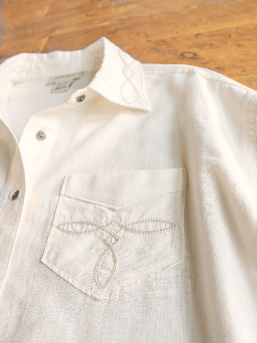 A close-up of The Lawler button-up top by Savannah Sevens western life{&}style, S-XXL, with an embroidered collar and decorative front pocket stitching, displayed on a wooden surface.