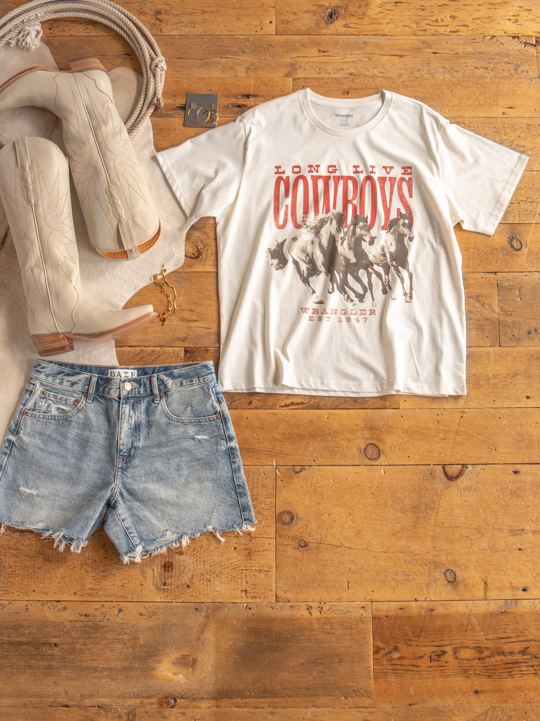 Flat lay of The Long Live graphic tee by Savannah Sevens western life{&}style, paired with denim shorts, beige cowboy boots, a coiled rope, and a silver belt buckle on a wooden floor.