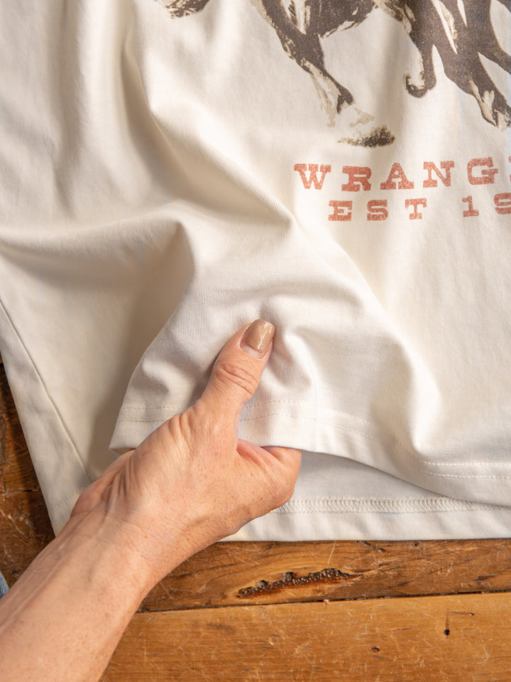 A hand pinches the hem of The Long Live white oversized tee by Savannah Sevens western life{&}style. Part of the shirt's graphic displays faded red letters. The tee rests on a wooden surface.