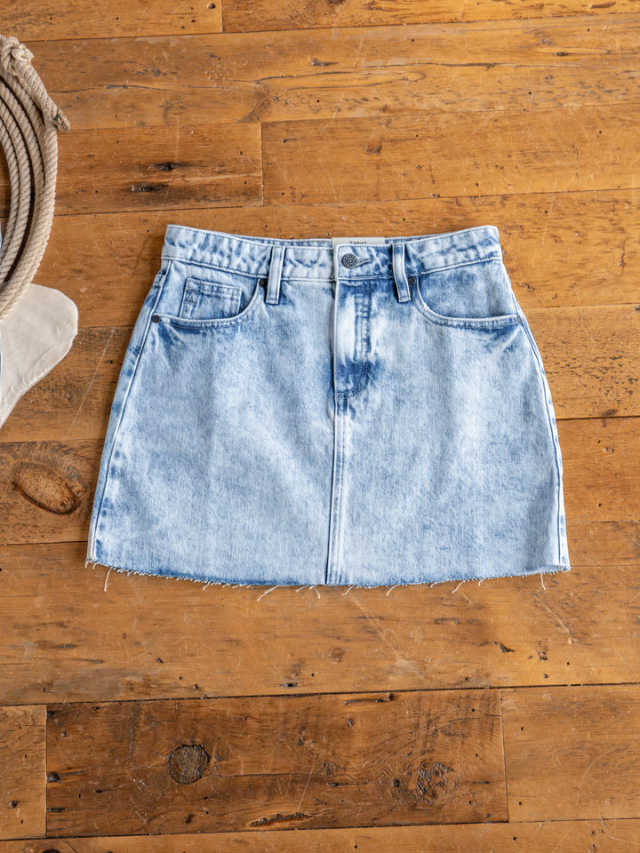 The Polson Skirt by Savannah Sevens western life{&}style, an acid wash denim mini with raw cut hem, front pockets, belt loops, and button closure, is laid flat on a rustic wooden floor beside a coiled rope.