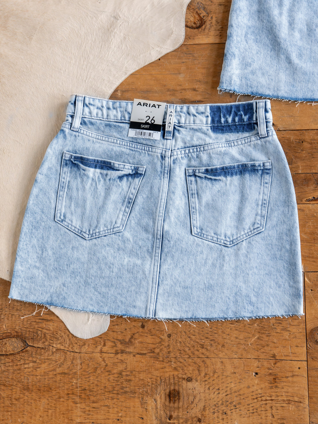 The Polson Skirt by Savannah Sevens western life{&}style, acid wash denim with raw cut hem and back pockets, is laid flat on a wooden floor with a tan cowhide rug partially visible underneath.