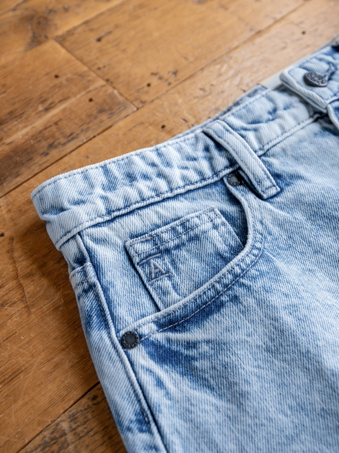 The Polson Skirt by Savannah Sevens western life{&}style is a light blue acid wash denim mini skirt with a small pocket, visible stitching, belt loops, and a raw cut hem, featuring a faded distressed look and shown on a wooden floor.