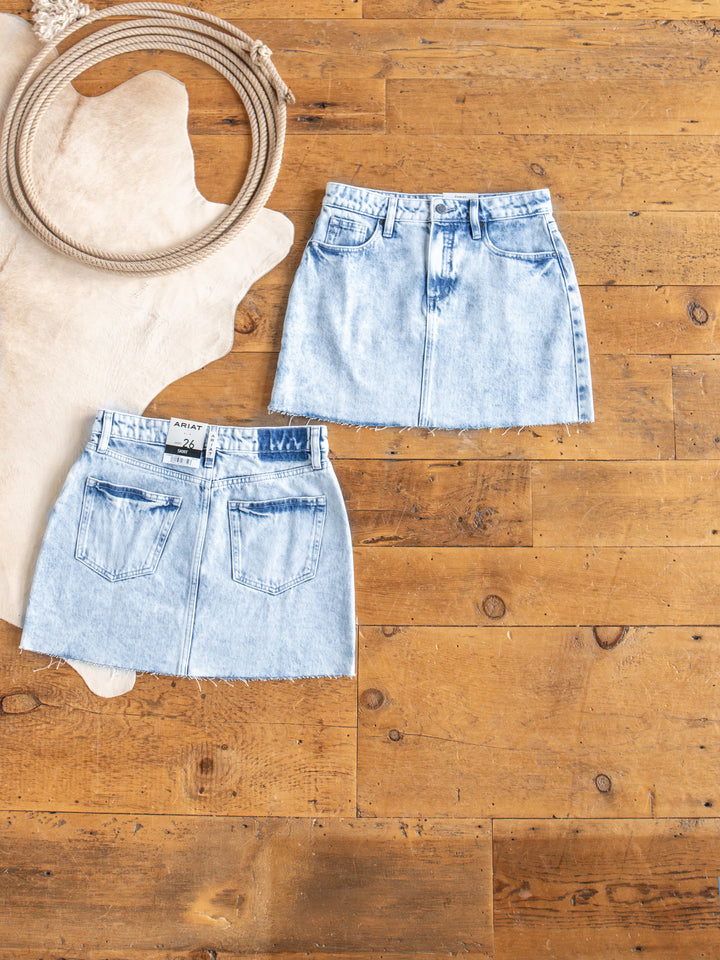Two Polson Skirts by Savannah Sevens western life{&}style, in light blue acid wash denim with raw cut hems, are displayed front and back on a wooden floor beside a coiled rope and a light animal hide rug.