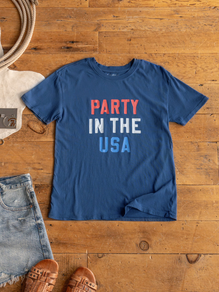 A blue "The Party" graphic tee by Savannah Sevens western life{&}style with red, white, and blue lettering is laid flat on a wooden floor next to denim shorts, brown sandals, and a beige tote bag.