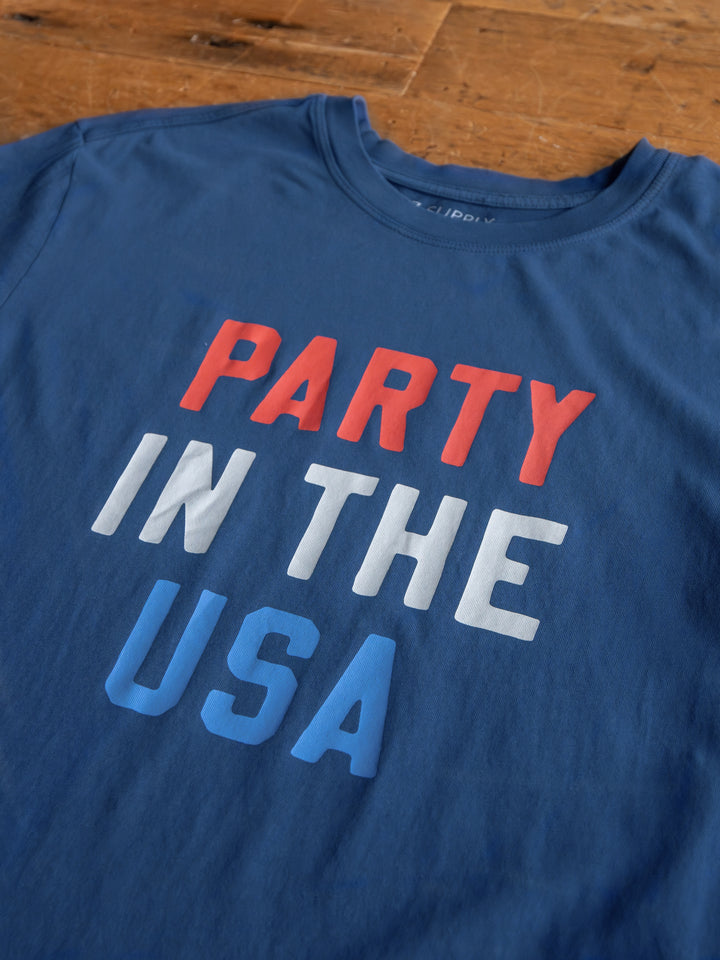 The Party tee by Savannah Sevens western life{&}style: a blue cotton shirt with "PARTY IN THE USA" in red, white, and blue lettering, displayed flat on a wooden surface.
