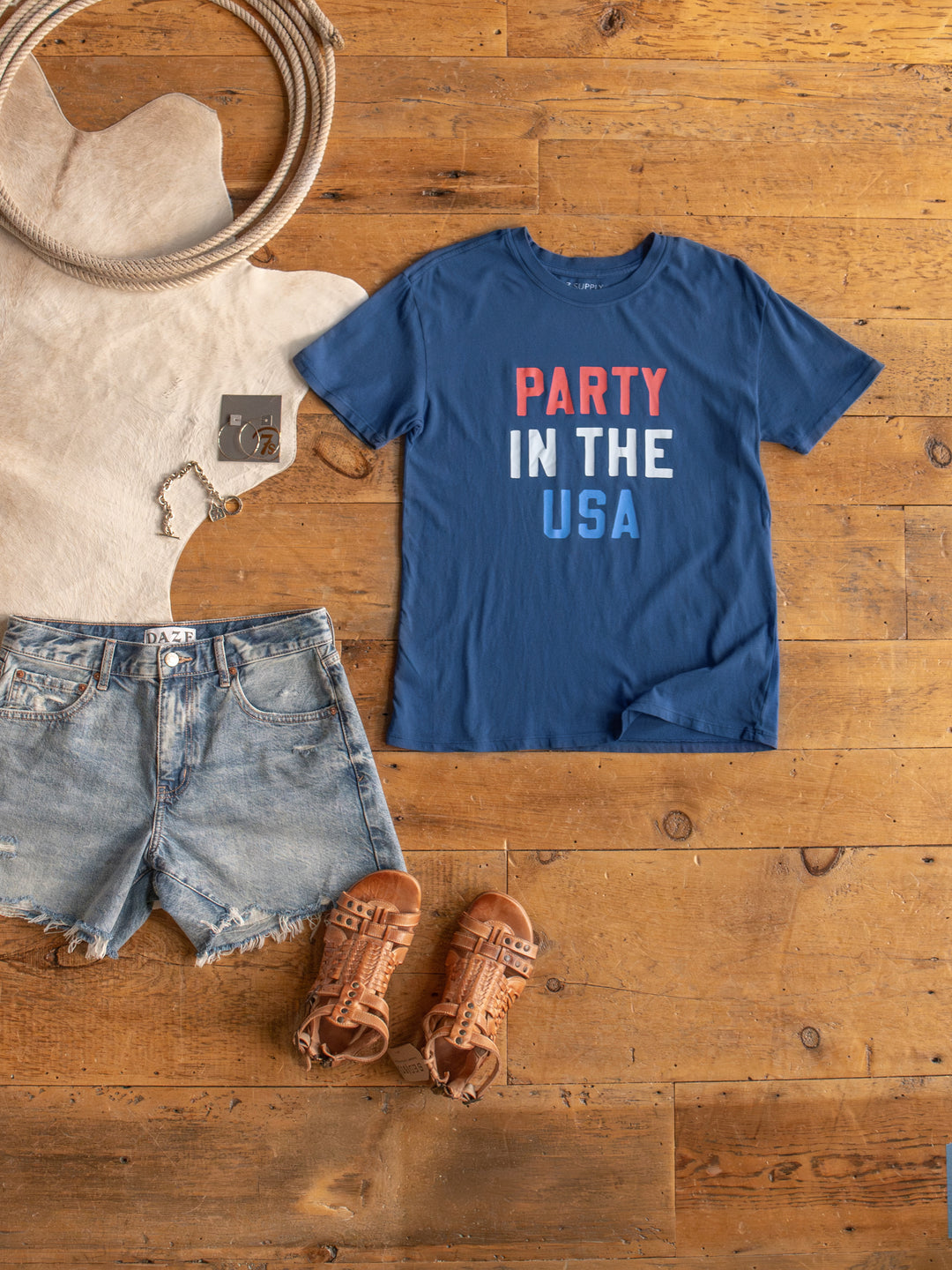 The Party tee by Savannah Sevens western life{&}style is a blue cotton graphic shirt with “PARTY IN THE USA” in red, white, and blue text, styled on a wooden floor with denim shorts, brown sandals, and a bracelet nearby.