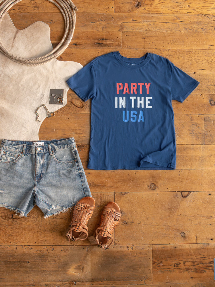The Party tee by Savannah Sevens western life{&}style is a blue cotton graphic shirt with “PARTY IN THE USA” in red, white, and blue text, styled on a wooden floor with denim shorts, brown sandals, and a bracelet nearby.