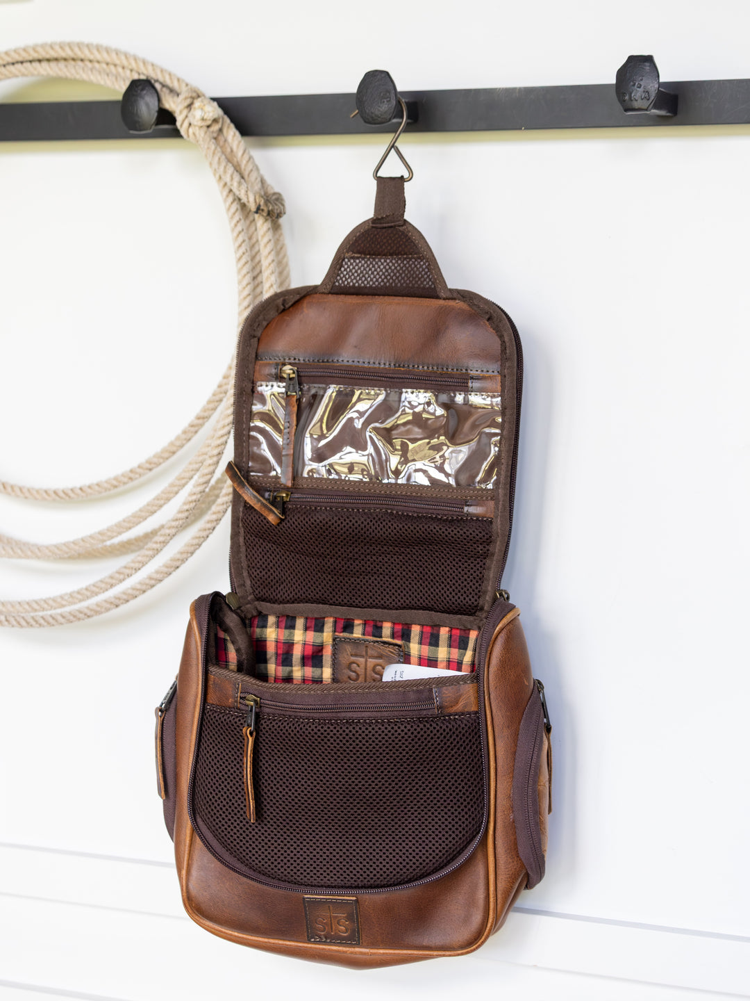 A Waynesboro Shave Kit by Savannah Sevens western life{&}style, featuring brown leather, mesh pockets, and plaid lining, hangs open from a hook on a white wall beside a coiled rope.