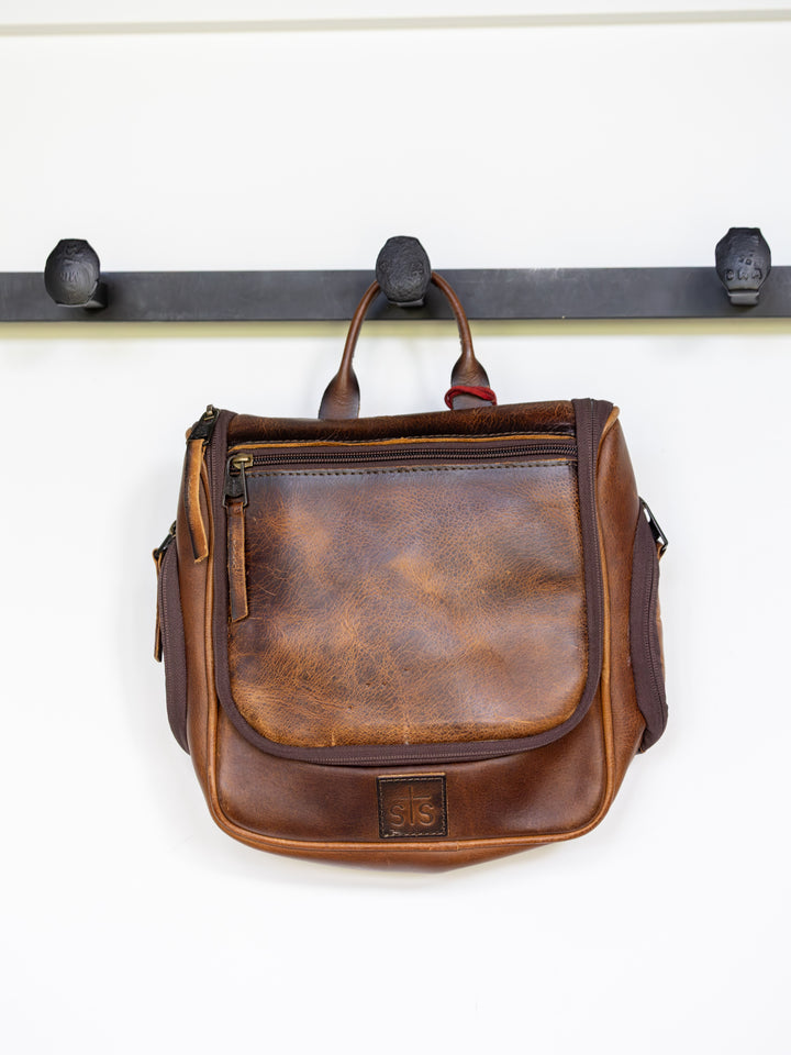 A brown leather Waynesboro Shave Kit by Savannah Sevens western life{&}style, featuring a front zipper pocket and top handle, hangs on a black three-hook wall-mounted rack, echoing a classic western look.