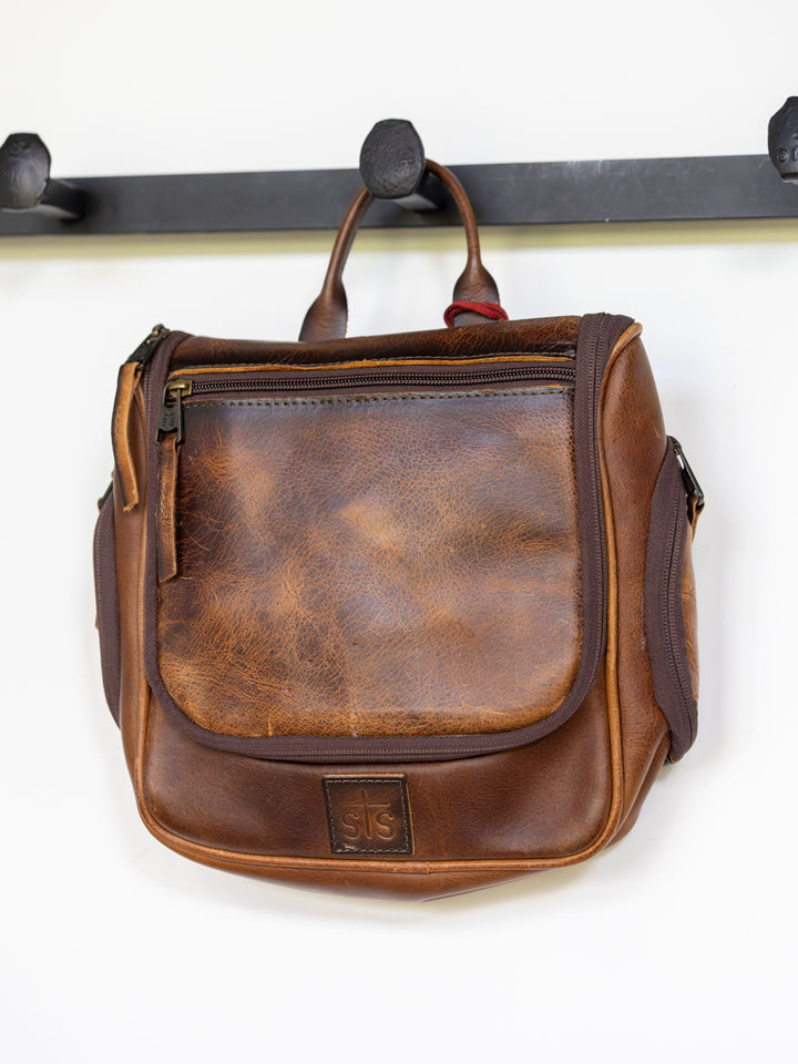 A brown Savannah Sevens western life{&}style Waynesboro Shave Kit with a front pocket, zipper, handle, and stitched logo patch hangs on a black wall hook. The leather displays natural wear.
