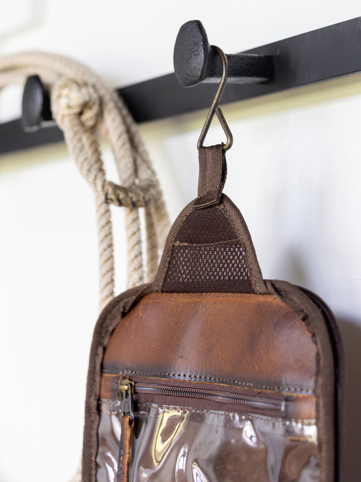 A brown leather and mesh Waynesboro Shave Kit by Savannah Sevens western life{&}style, featuring a clear plastic front and zipper pocket, hangs on a black rack. Coiled rope is on the next hook.
