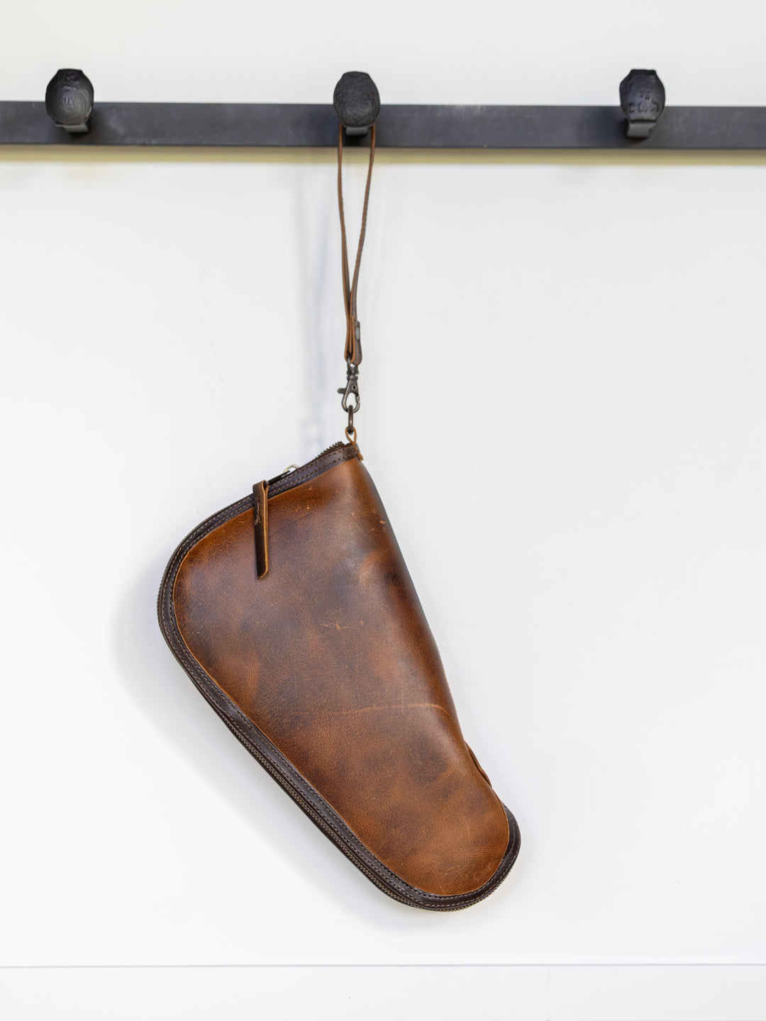 The Waynesboro Pistol Case by Savannah Sevens western life{&}style with a zipper and strap hangs from a black metal coat hook against a white wall.