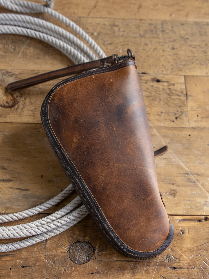 The Waynesboro Pistol Case by Savannah Sevens western life{&}style, crafted from brown full grain leather with visible wear, rests on a rustic wooden surface beside a coiled white rope.