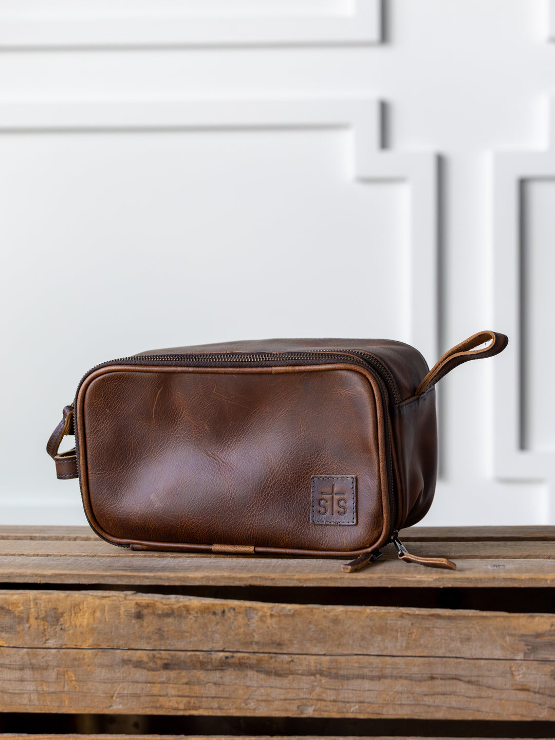 The Wayneboro Shave Bag by Savannah Sevens western life{&}style, crafted from full grain leather with a handle, sits on a rustic wooden surface against a white paneled background.