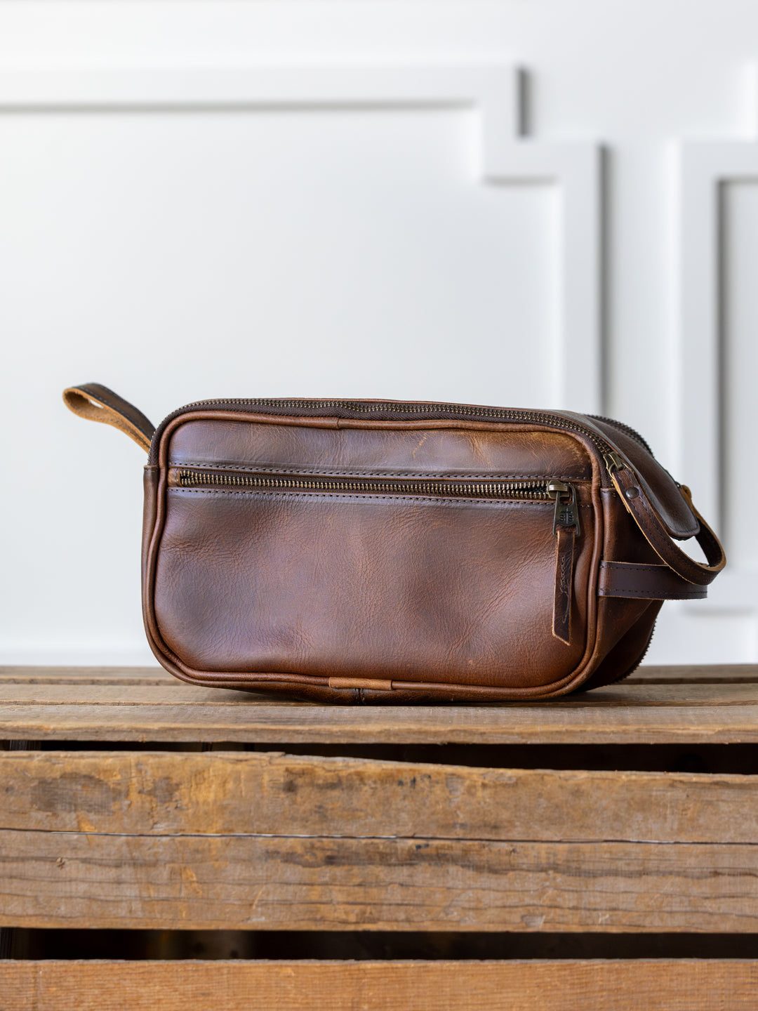 The Wayneboro Shave Bag from Savannah Sevens western life{&}style, made of full grain leather with a front zipper pocket, rests on a rustic wooden surface against a white paneled wall.