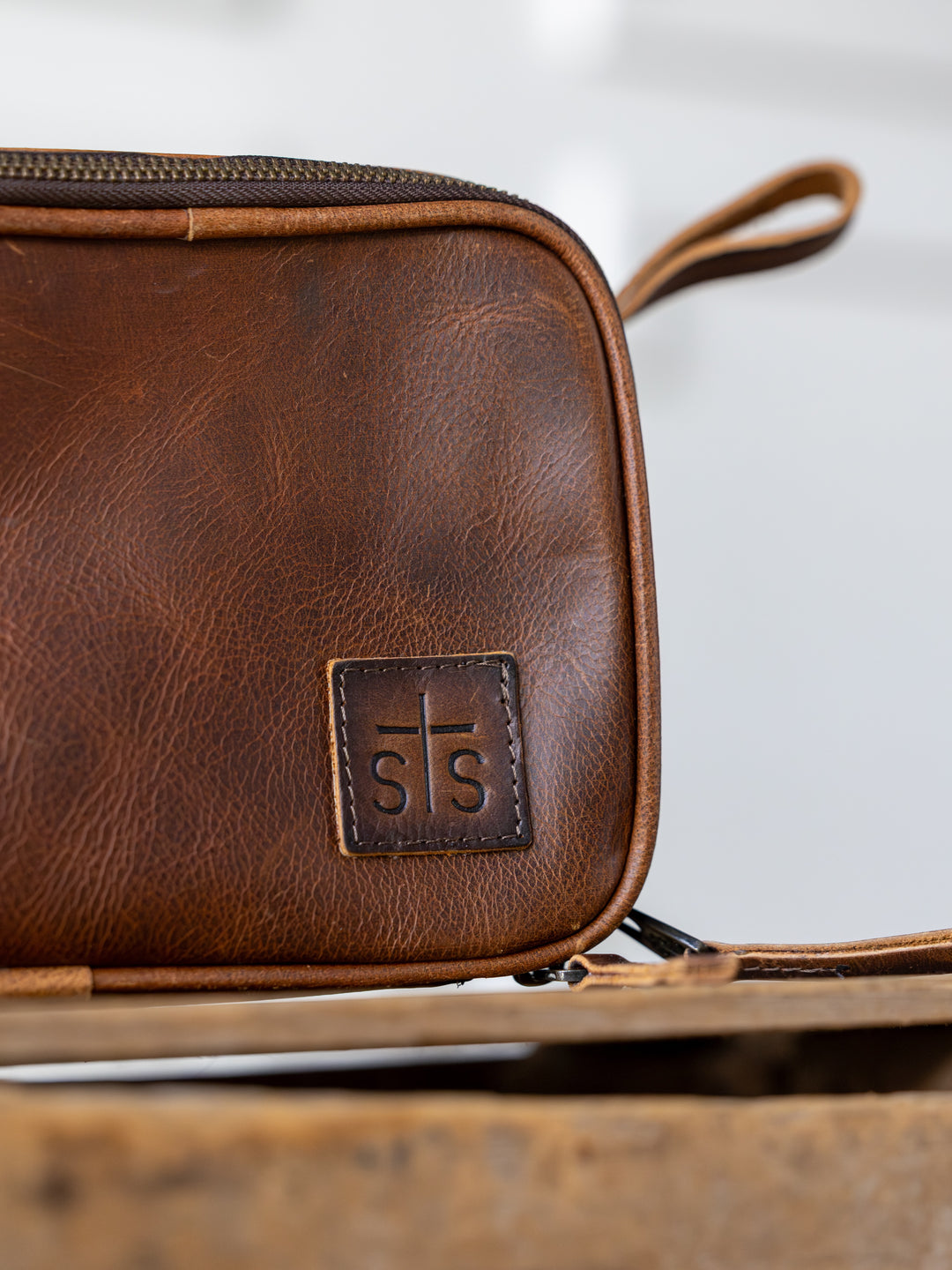 A close-up of the Wayneboro Shave Bag by Savannah Sevens western life{&}style, made of full grain leather with a stitched “s|s” patch, double zippers, and a handle, shown on a wooden surface with a blurred background.