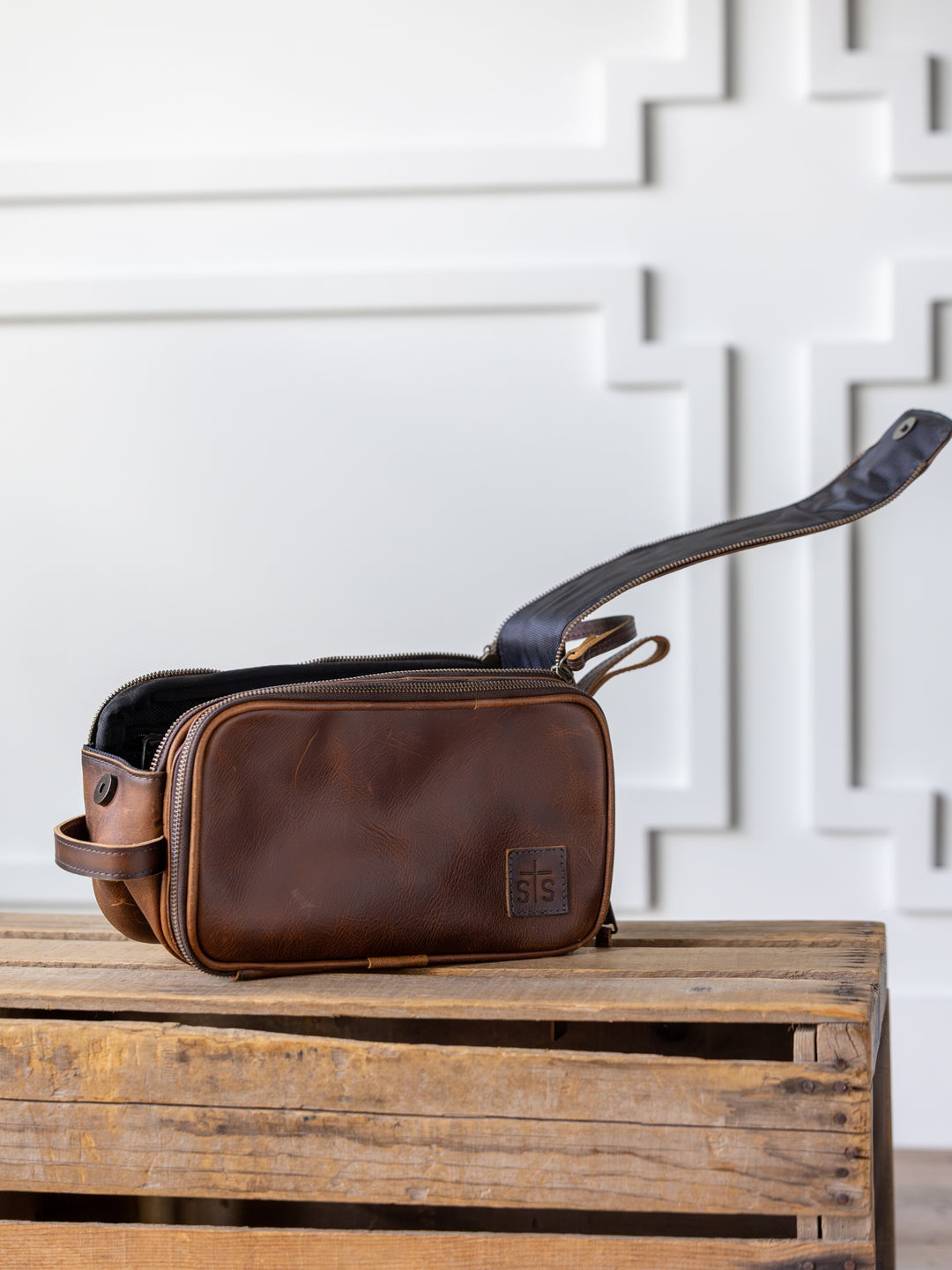 The Wayneboro Shave Bag by Savannah Sevens western life{&}style, crafted from full grain leather with a black zipper, sits open on a wooden crate against a white geometric-patterned wall.