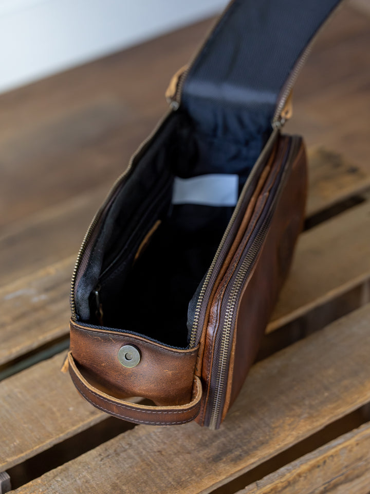 The Wayneboro Shave Bag by Savannah Sevens western life{&}style, crafted from full grain leather, is unzipped to show its black interior lining as it rests on a wooden surface.