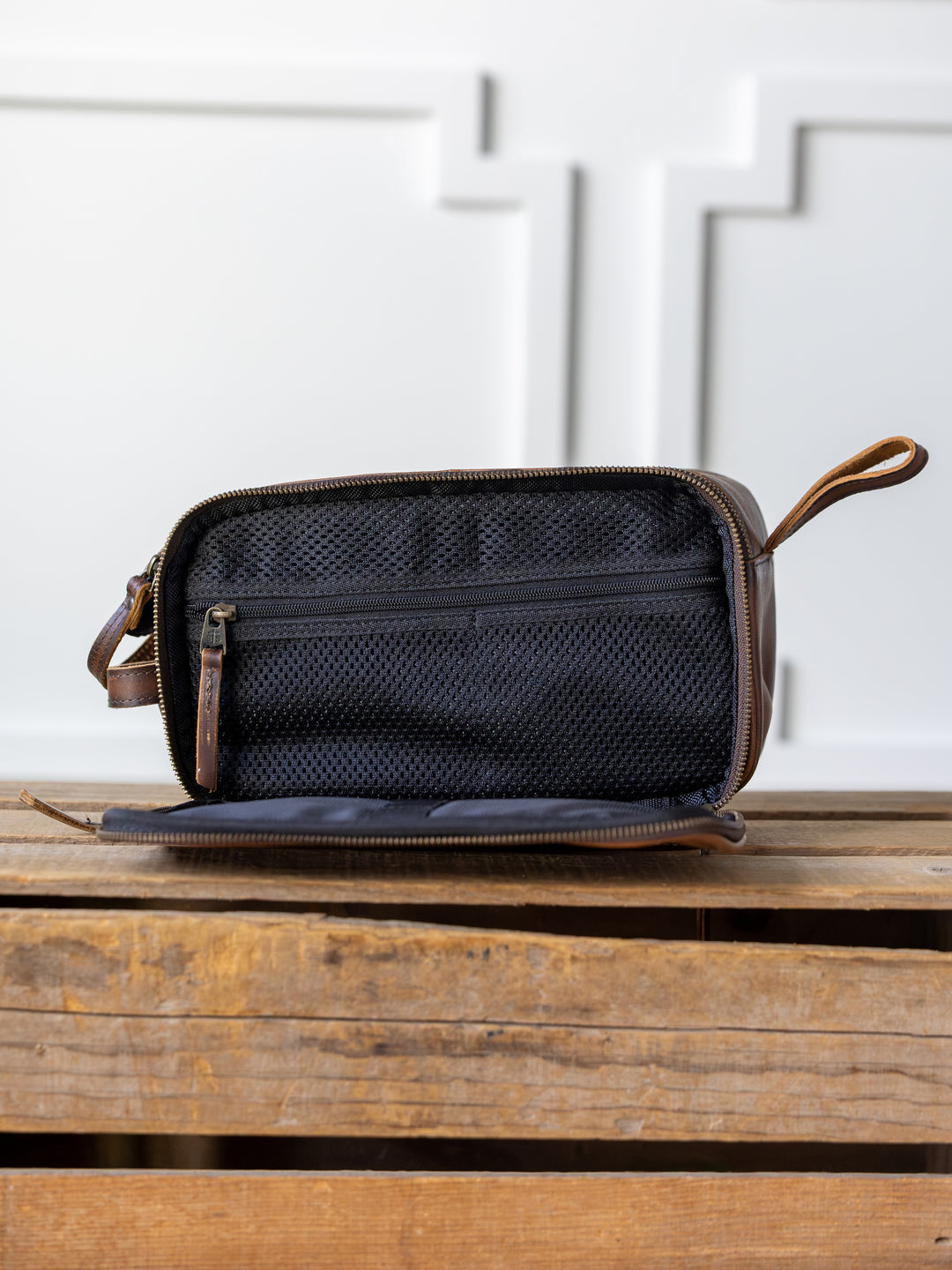 The Wayneboro Shave Bag by Savannah Sevens western life{&}style features brown full grain leather and a zipper, shown open to display a black mesh pocket inside. The bag sits on a wooden surface with a white textured wall behind it.