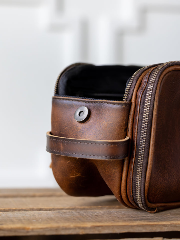 A close-up of the Wayneboro Shave Bag by Savannah Sevens western life{&}style, crafted from full grain leather with a zipper and buttoned strap, sits on a wooden surface against a blurred white background.