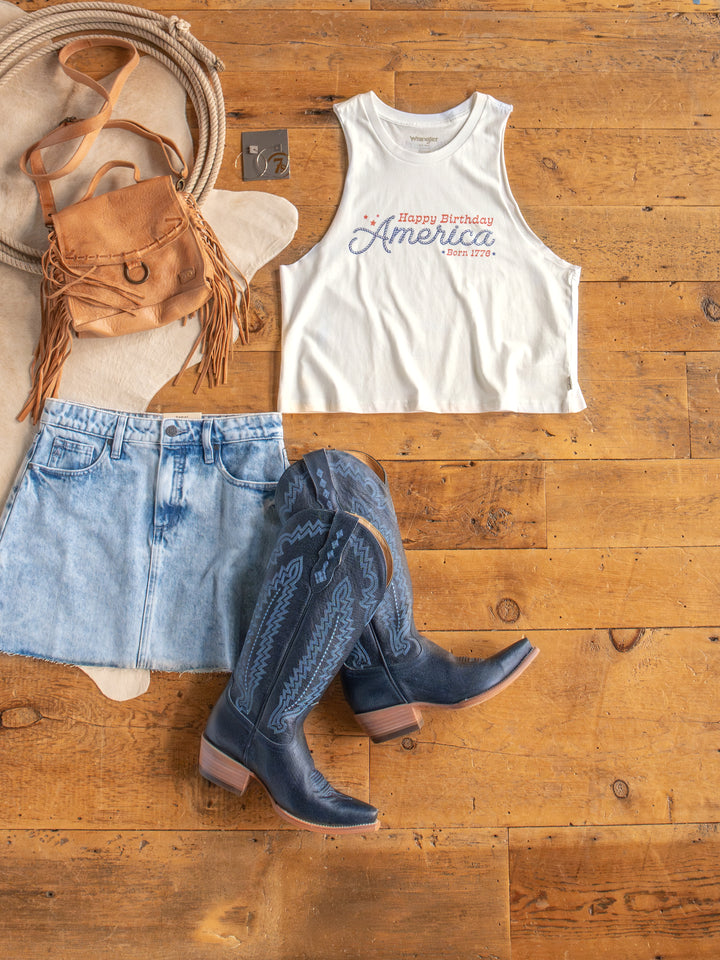 Flat lay of The HBD America muscle tank by Savannah Sevens western life{&}style, paired with a light denim skirt, blue cowboy boots, brown fringe bag, coiled rope, and a metal star on a wooden floor.