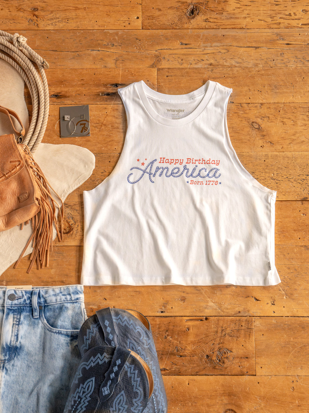 The HBD America tank top by Savannah Sevens western life{&}style is a white 100% cotton shirt with "Happy Birthday America Born 1776" in red and blue text, styled with denim shorts, blue sandals, a tan hat, and accessories.