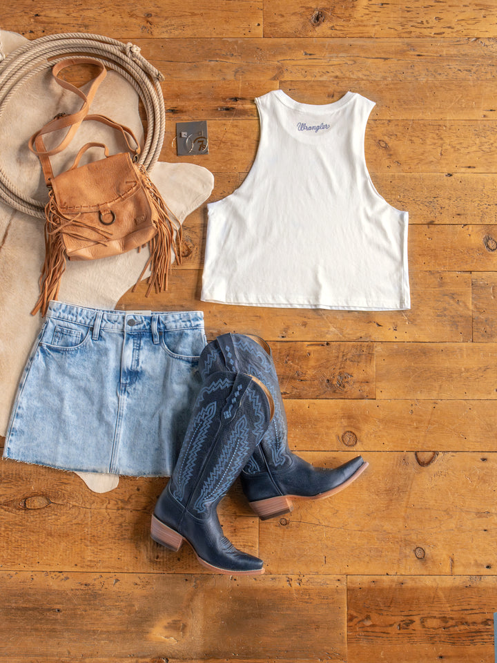 Flat lay of The HBD America muscle tank crop top by Savannah Sevens western life{&}style, paired with a light denim mini skirt, blue cowboy boots, tan fringed leather bag, and silver earrings on a wooden floor.