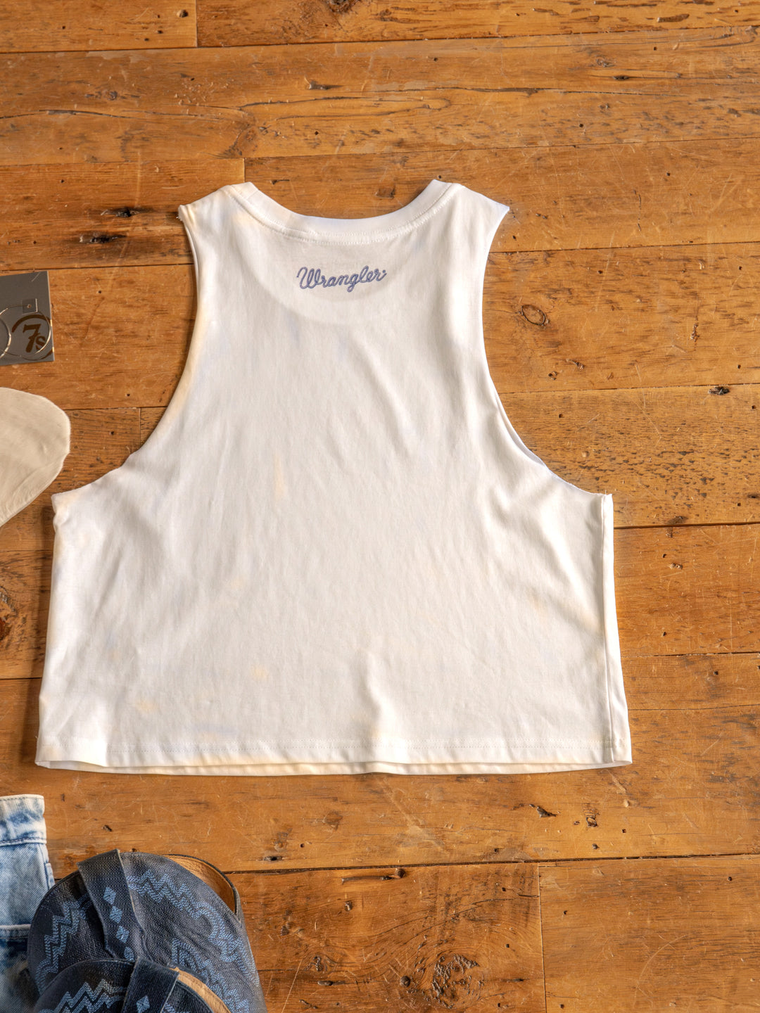 The HBD America by Savannah Sevens western life{&}style is a sleeveless white muscle tank made from 100% cotton, laid flat on wood with denim and blue patterned fabric peeking out—perfect for celebrating Happy Birthday America.