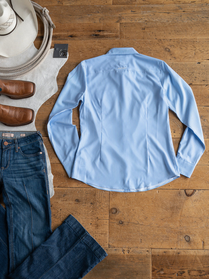 The Ellisville by Savannah Sevens western life{&}style—a light blue pinstripe, long-sleeve sun shirt—lies backside up on a wooden floor with a cowboy hat, brown boots, and blue jeans arranged nearby.