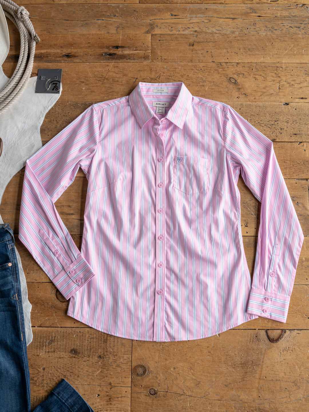 The Kinnear by Savannah Sevens western life{&}style, a pink and white striped long sleeve top, is displayed on a wooden floor next to dark blue jeans and a coiled rope.