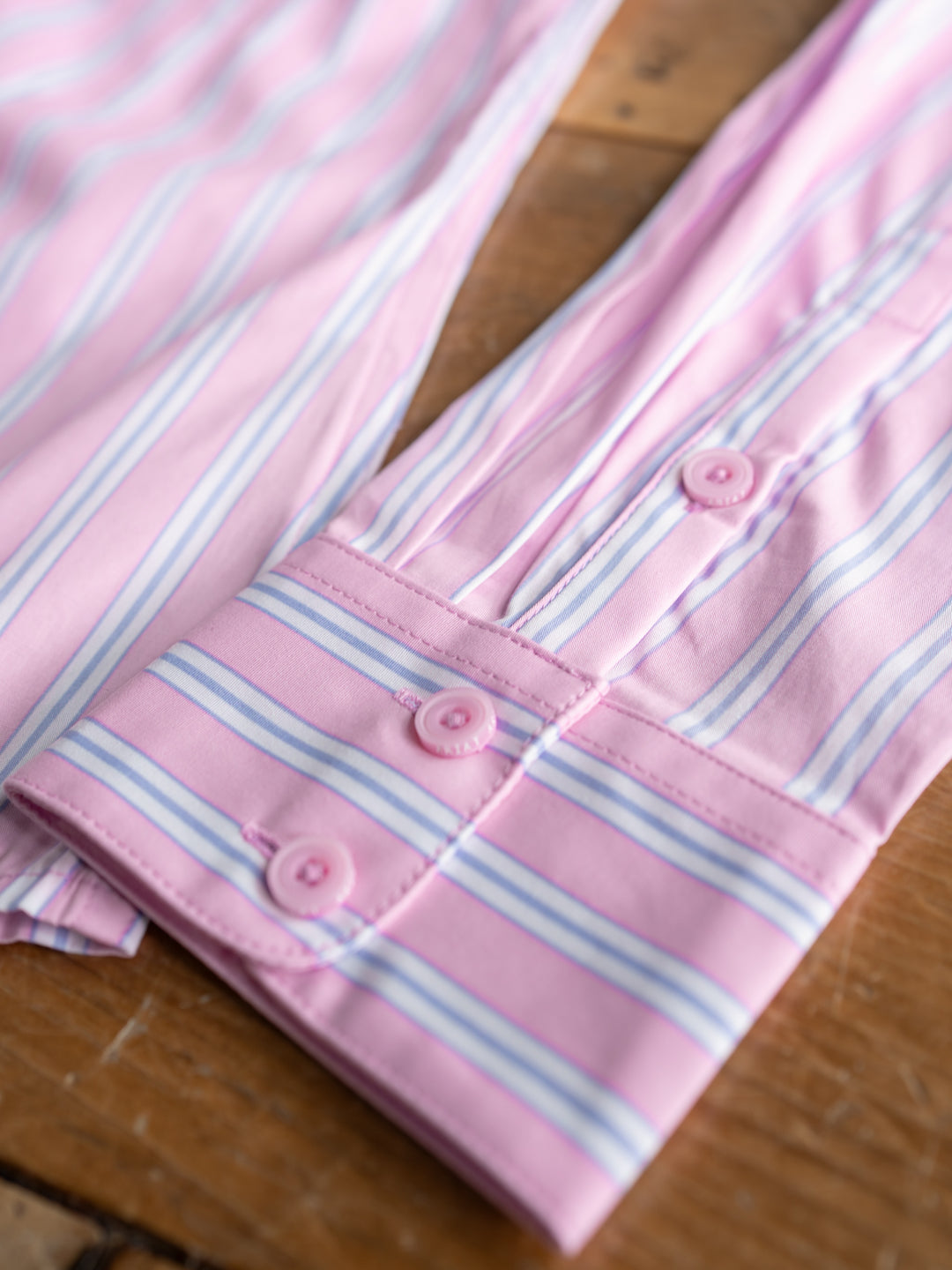 A close-up of The Kinnear by Savannah Sevens western life{&}style, a fitted striped long sleeve top in pink, light blue, and white with three pink cuff buttons, laid flat on a wooden surface for a polished look.