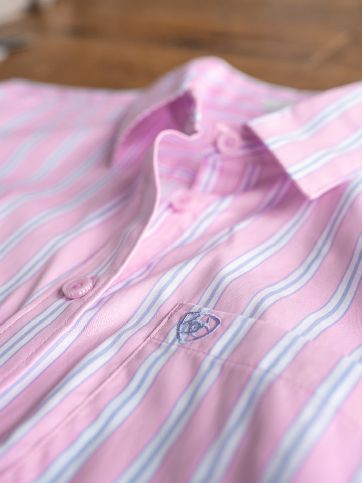 A close-up of The Kinnear by Savannah Sevens western life{&}style, a wrinkle-resistant pink long sleeve top with blue and white stripes, buttons, and an embroidered chest logo, neatly folded on a wooden surface.