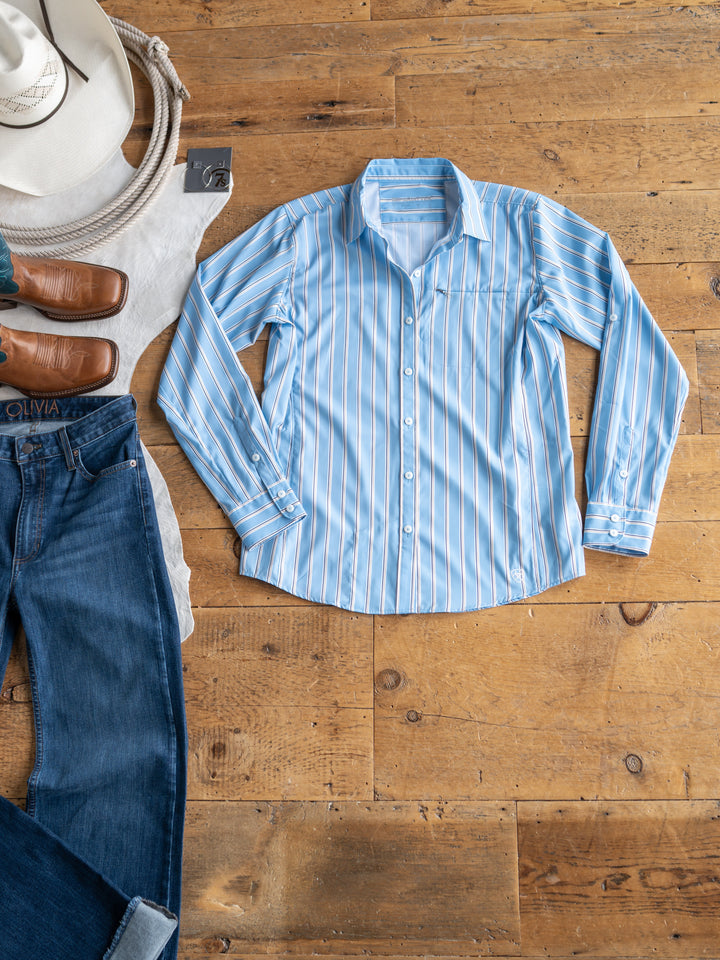 A light blue and white striped The Colmar long sleeve top by Savannah Sevens western life{&}style is neatly laid out on a wooden floor with blue jeans, brown boots, a white cowboy hat, a belt, and a coiled rope. This sun shirt is perfect for any adventure.