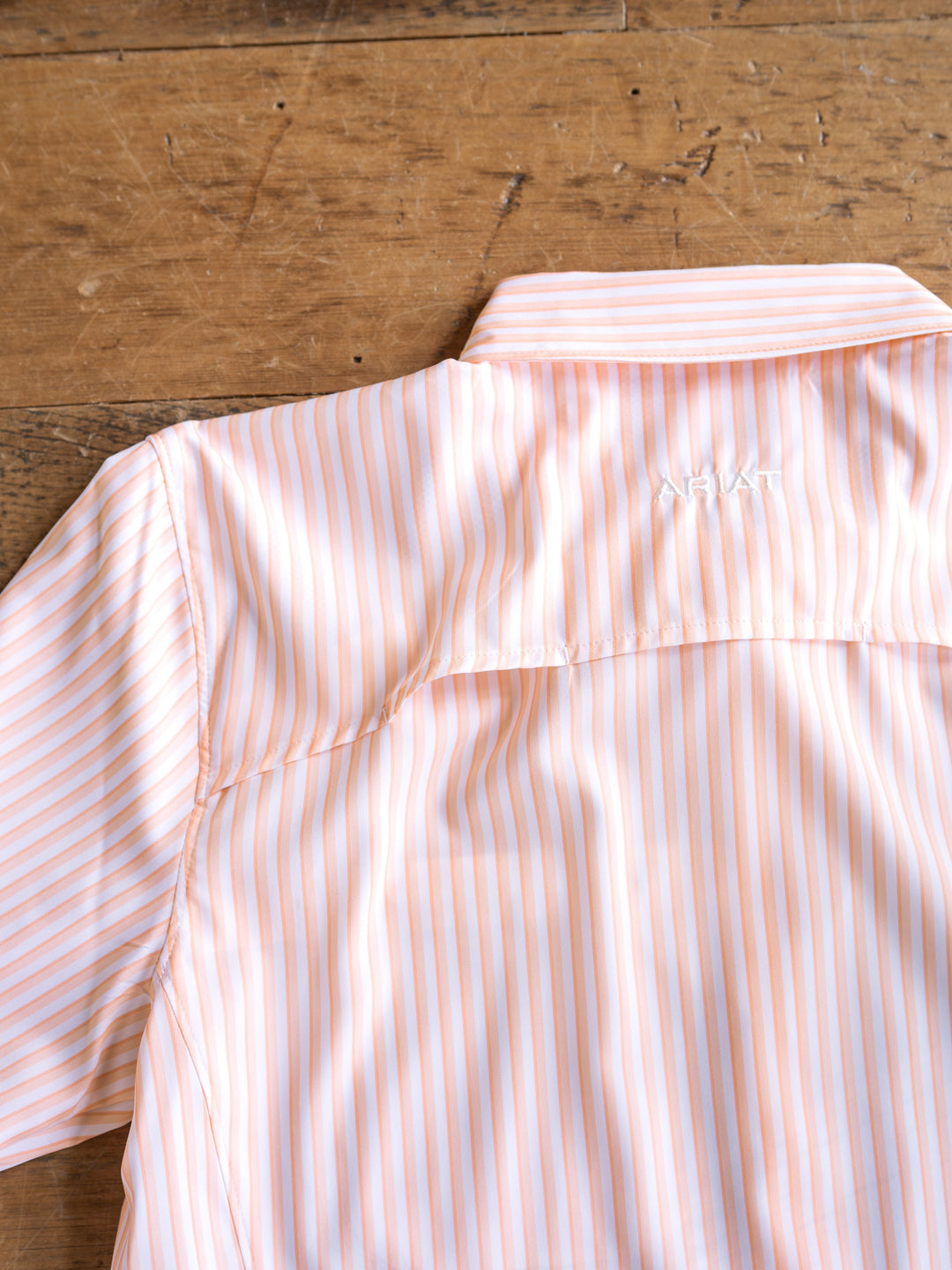 The Lovilia by Savannah Sevens western life{&}style is a light orange and white striped long sleeve shirt with a collar, featuring moisture wicking, sun protection, and "ARIAT" embroidered in white on the back shoulder.