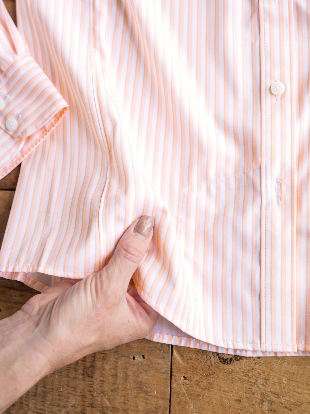 A hand holds the bottom edge of The Lovilia by Savannah Sevens western life{&}style, displaying its striped long sleeves and buttons. This sun protection shirt is laid flat on a wooden surface.
