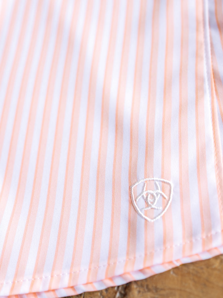 Close-up of The Lovilia by Savannah Sevens western life{&}style, a striped long sleeve top with light pink and white vertical stripes and a small white embroidered shield logo near the hem. Moisture-wicking fabric keeps you comfortable all day.