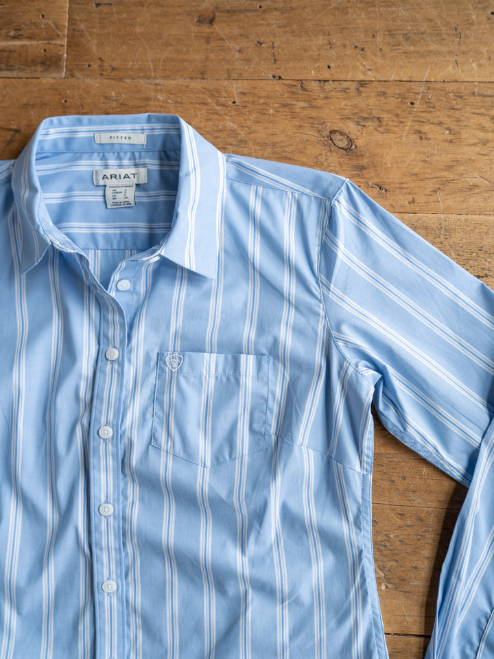 The Ingall by Savannah Sevens western life{&}style is a light blue fitted long sleeve button-up with white vertical stripes, a left front pocket, and an “ARIAT” label on the collar, shown laid flat on a wooden surface.