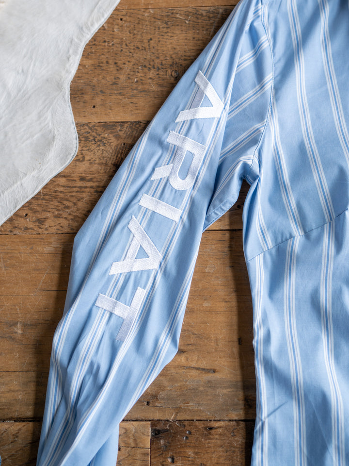 The Ingall by Savannah Sevens western life{&}style is a light blue, striped fitted long sleeve button-up with wrinkle-resistant fabric and "ARIAT" in white on the sleeve, shown on wood beside a white cloth. Available in sizes S-XXL.