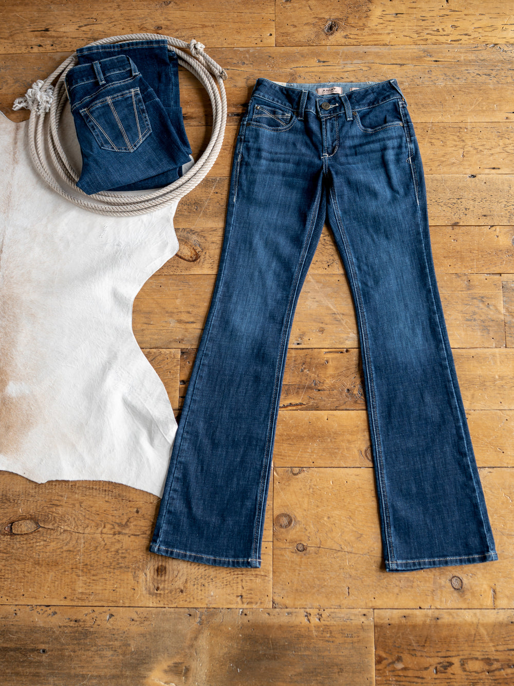 The Avery Perfect Rise Boot Cut Jean by Savannah Sevens western life{&}style in medium dark wash is laid flat on a wooden floor beside a coiled rope, folded jeans, and a white cowhide rug.
