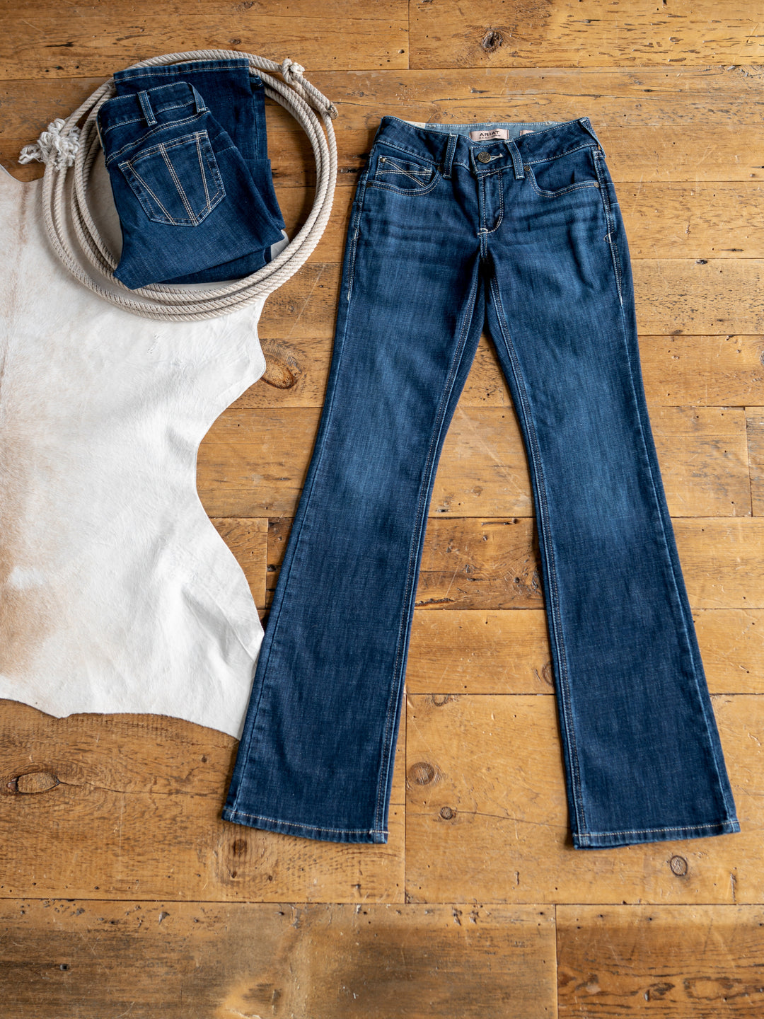 The Avery Perfect Rise Boot Cut Jean by Savannah Sevens western life{&}style in medium dark wash is laid flat on a wooden floor beside a coiled rope, folded jeans, and a white cowhide rug.