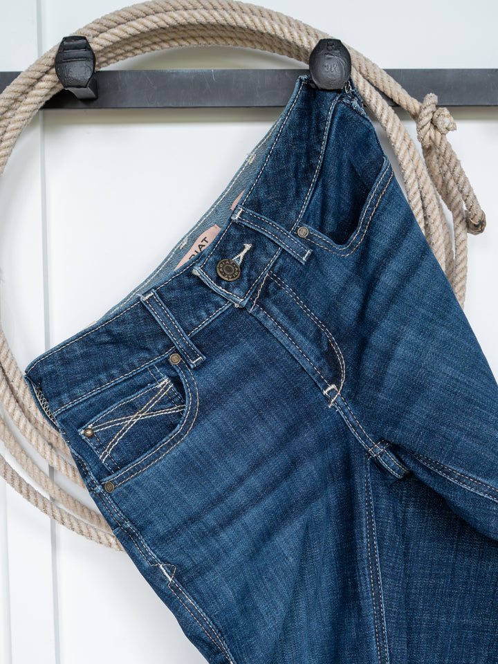 The Avery Perfect Rise Boot Cut Jean by Savannah Sevens western life{&}style in medium dark wash hangs by a belt loop on a black hook, with a coiled beige rope behind it against a white background.