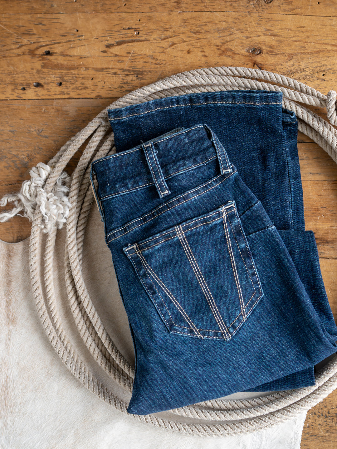 The Avery Perfect Rise Boot Cut Jean by Savannah Sevens western life{&}style, in a medium dark wash, sits atop a coiled rope on wood, with a touch of cowhide peeking from the corner.