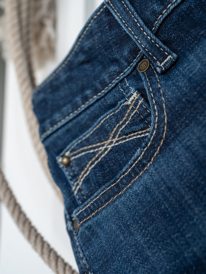 Close-up of the front pocket and belt loop of The Avery Perfect Rise Boot Cut Jean by Savannah Sevens western life{&}style in a medium dark wash with visible stitching and rivets, with a blurred rope in the background.