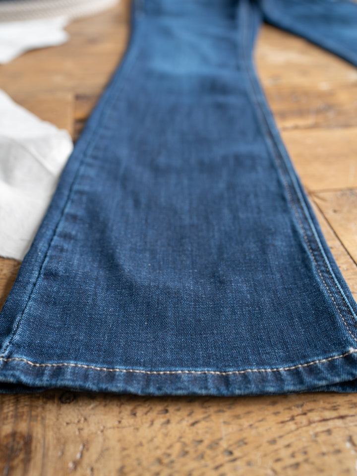A close-up of The Avery Perfect Rise Boot Cut Jean by Savannah Sevens western life{&}style in a medium dark wash with flared bottoms, laid flat on a wooden surface.